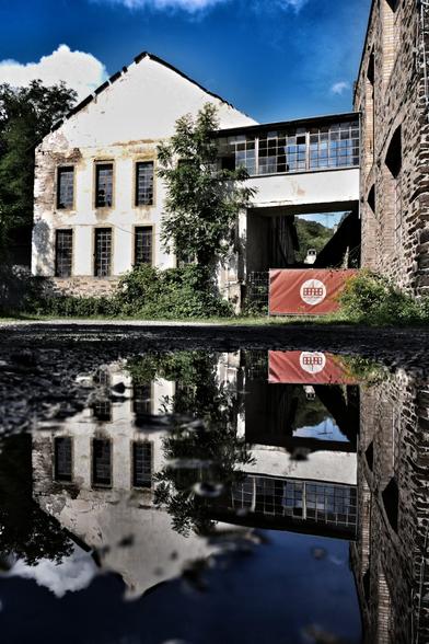 Eine marode, stillgelegte Wollfabrik (zu lesen auf einer roten Folie, die an einer Absperrung angebracht ist) spiegelt sich in einer Pfütze. Zwei Gebäudeteile sind durch einen Durchgang im 1. Stock verbunden. Viele der Glasfenster sind kaputt. Vor dem Gebäude wachsen Bäume und Sträucher.
