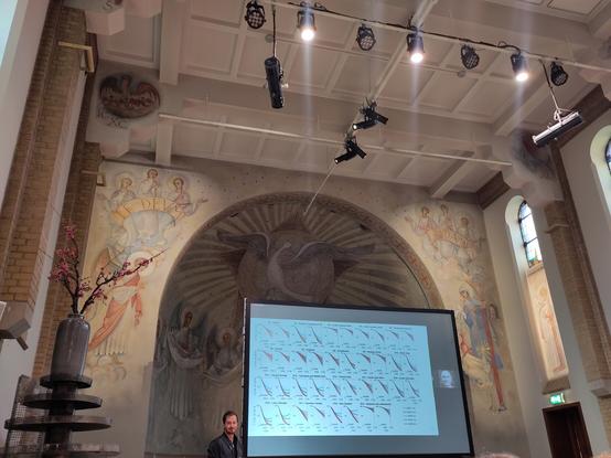 A man stands in front of a screen showing many small graphs with red lines all pointing downwards. Behinds a painted wall with a curved alcove. A dove of peace is in the centr and groups of Angels on aleiter side hold banners with the texts Te Deum and Laudamus painted on