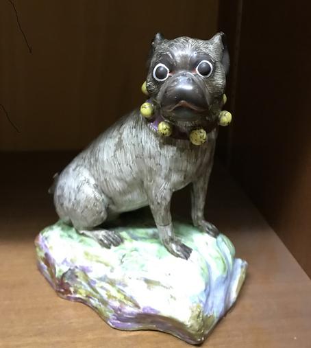 eine Figur aus Stein: der Mops mit einem gelben Perlen-Halsband sitzt auf einem Stein in einem dunklen Schrank