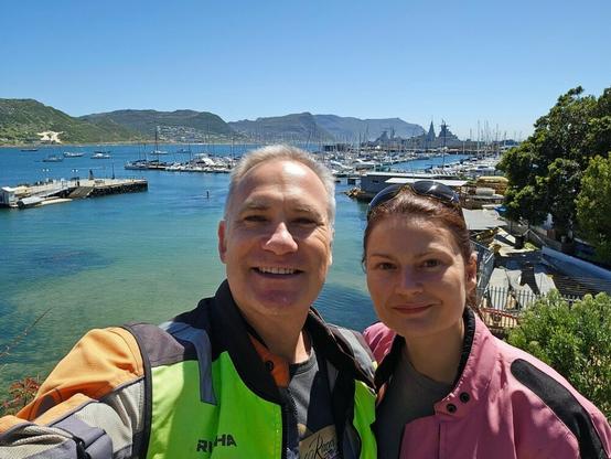 Man and a woman smiling at the camera, with a harbour in the background with lots of yachts moored at the jetty.