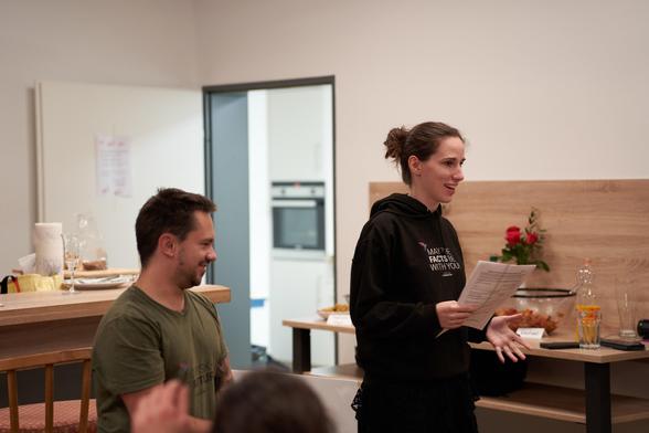 Ich stehe strahlend in einem schwarzen Hoodie im Vordergrund und halte eine Rede. Links neben mir steht ein kleinerer Mann, der ebenfalls lacht. Im Hintergrund sieht man eine Bar und den Eingang zu einer Küche.