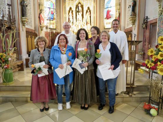 Blick in den erleuchteten Altarraum einer kleinen neugotischen Kirche: Vorne stehen vier lachende und lächelnde Frauen, die jede eine Rose und eine Urkunde in Händen halten, dahinter zwei Männer in Albe und eine weitere Frau, die ebenfalls gut gelaunt dreinschauen. Noch dahinter sind ein glänzender geschnitzter Hochaltar und Buntglasfenster zu erkennen, rechts und links sind Blumenschmuck und Erntegaben zu sehen.