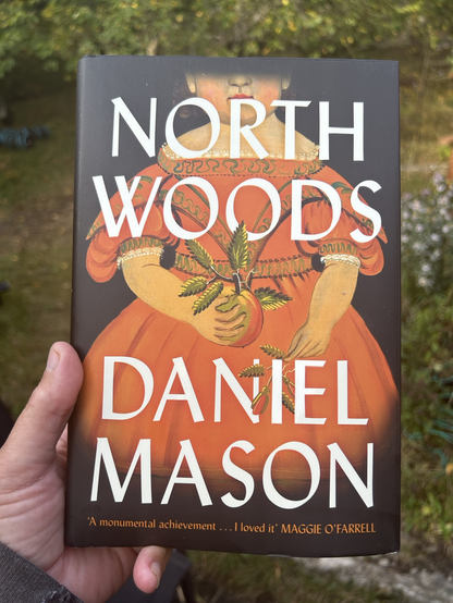 Closeup of the cover of Northern Woods by Daniel Mason. There's also a blurb on the cover, by Maggie O'Farrell that reads: 'A monumental achievement ... I loved it'. The cover illustration is a painting or drawing of a long girl in an old-fashioned orange dress. In her right hand she holds an apple, with stem and leaves attached. In her left hand she loosely holdd similar leaves. The dress and the girls's bare lower arms (plus apple) take up most of the page. Only the lower half of the girl's face is in the frame, and even the bottom of the dress is out of the frame. Of the girl's face only the mouth and the lower part of her nose and ears are visible.