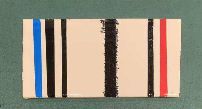 Off-white rectangular tile with stripes. A blue stripe followed by two black stripes on the left side, a black stripe followed by a red stripe on the right side, and a thick black stripe with small florets of black coming out the side in the middle
