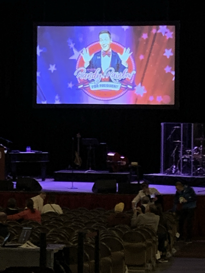 The stage before the show. Black and lit dark purple with piano, drum kit, and other instruments. The screen behind is lit with the logo for the show