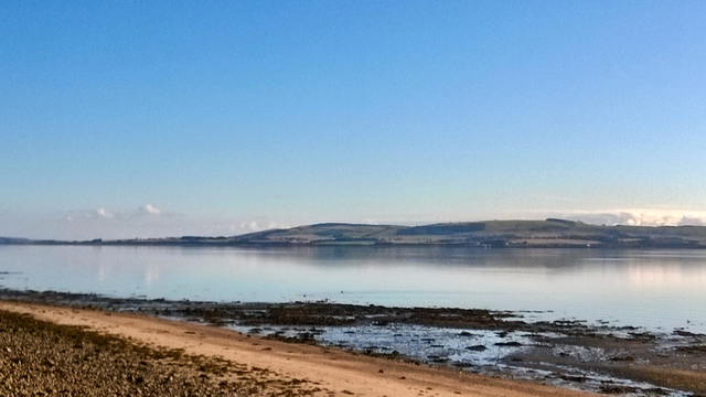 A photo of a calm sea, the Cromarty Firth at Salt urn, looking to the Blck Isle. A pale blue sky, with a thin layer of trite cloud, slightly tinted pink. A double hill profile on the far shore, horizon level, showing fields and teees, a slight reflection. Stock still water. Some seaweed nd a sandy foreshore .