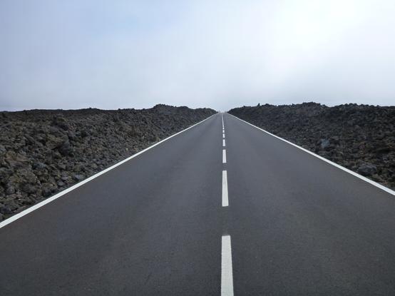 Route rectiligne à deux voies, bordée de coulées de lave issue du volcan Teide sur l'ile de Tenerife, aux Canaries. De la brume masque tout le dessus de l'horizon.