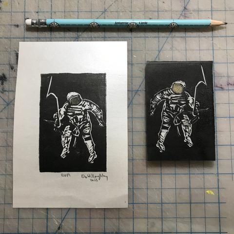 Tiny astronaut on a spacewalk linocut printed in black ink with a hint of gold on the visor, on shiny white paper with a pearlescent sheen, next to the 2” x 3.5” inked block, on a cutting mat with a half-inch grid with a pencil for scale.