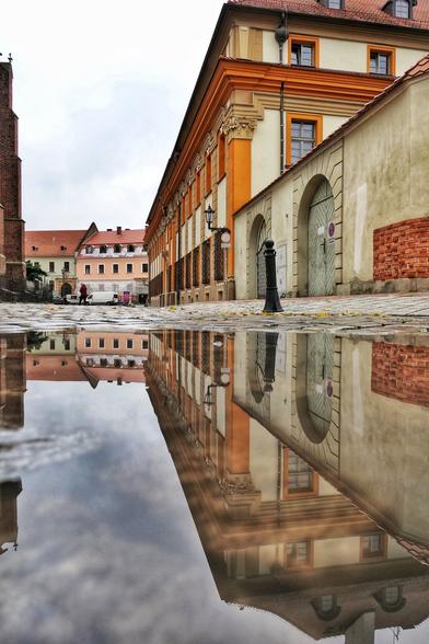 In einer Pfütze auf einer Kopfsteinpflaster-Straße spiegeln sich das rechts angrenzende historische Gebäude (hell-beige mit orangefarbenen eckigen Säulen und Fensterumrandungen), sowie eine Mauer mit zwei gewölbten, verschlossenen Toren. Davor ist ein schwarzer Poller zu sehen. Die Straße führt auf ein rosanes und ein grünes Haus zu, die sich auch in der Pfütze spiegeln. Vor diesen Häusern stehen mehrere Autos, davon ist eins ein weißer Transporter. Eine Person überquert vor dem Häusern die Straße. Der Himmel ist bewölkt.