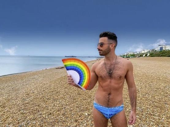 shirtless white gay man with dark hair, moustache, stubble and sunglasses, looking sideways, wearing blue speedos and holding a rainbow fan, on a gravel-sand beach in Brighton