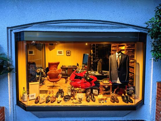 A blue wall and a glass bay window of a leather shoe store at dusk. The interior of the store is lit in yellow lights and there are many shoes on the inside wooden ledge. There is a suit and tie dressed on a headless mannequin and a leather chair inside the store. A red sports bike is also inside the store