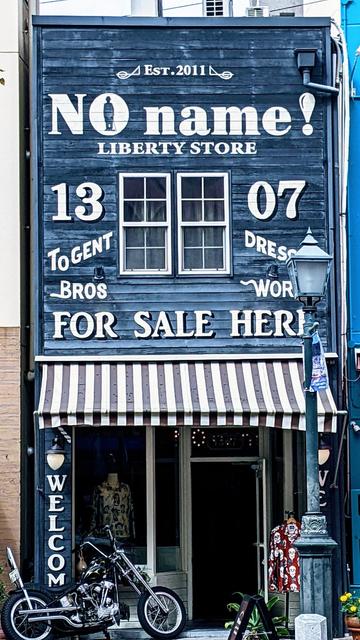 A storefront on two floors with the upper storey outside with blue painted wood and white trimmed window and large white lettering that notes est 2011 No Name !I erty Store 
And below to gent bros, and dress work
And for sale here.
A motorcycle is parked outside the store