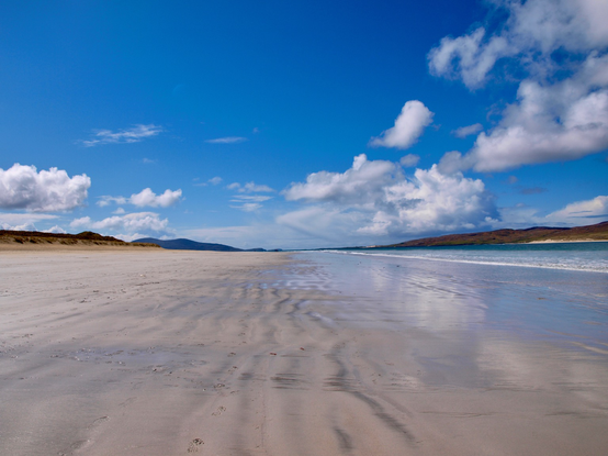 Beach Photo. A bright blue sky with puffy white cloud. Hills on the horizon, with a centre point where sea and shoreline meet. Sand across the expanse of the photo with the sea a triangle to the right. Cloud reflections in the wet sand.