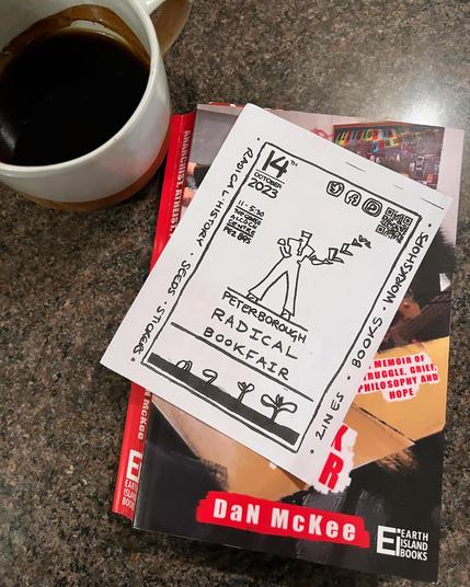 A cup of coffee next to a copy of DaN McKee’s book, Anarchist Atheist Punk Rock Teacher with a flier for the Peterborough Radical Bookfair (11am-5.30pm at the George Alcock Centre, Saturday October 14th) resting on top.