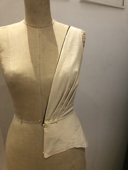 Front view of a dress form with muslin draped into a plunging neckline with pleats at the waist and a peplum