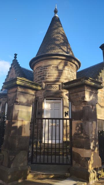 Small, stone gatehouse, with crow step roof ends, and small, turret style tower entrance with 1863 inscribed above doorway, seen framed between two stone gate pillars