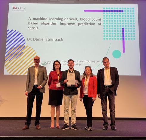 5 persons on stage, in the middle one honored man holding a price. In the background there is a powerpoint slide showing the name of the prizewinning scientific work "A machine learning-derived, blood count based algorithm improves prediction of sepsis. Dr. Daniel Steinbach"