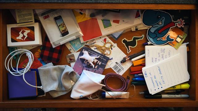 A desk drawer with stamps, measuring tape, twine, cleaning cloths, stickers, pens, pencils, to-do lists, a patch, notebook, old iPhone and scissors