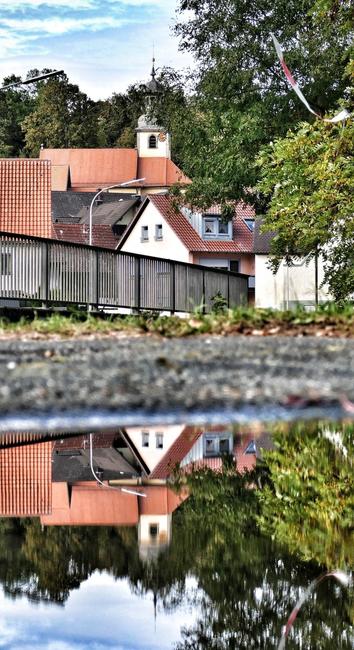 Das Bild zeigt ein kleines Dorf mit einer Kirche und Häusern. Das Bild ist horizontal in der Mitte geteilt, wobei die obere Hälfte das Dorf und die untere Hälfte eine Spiegelung des Dorfes in einem Gewässer zeigt. Das Dorf besteht aus einer Kirche mit einem weißen Turm und rotem Dach sowie mehreren Häusern mit roten Dächern. Die Häuser sind von Bäumen und Sträuchern umgeben. Die Spiegelung im Wasser ist fast ein perfektes Spiegelbild des Dorfes. Der Himmel ist blau mit einigen Wolken.