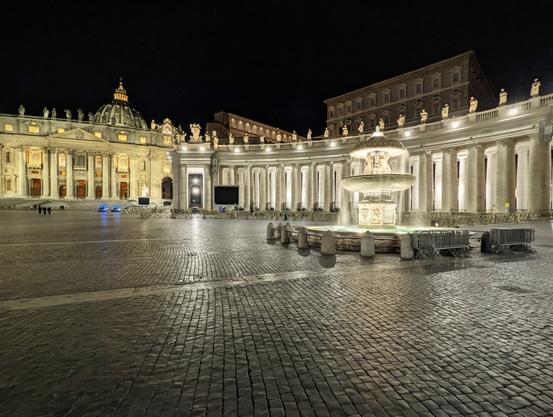This is a nighttime photo of a large cobblestoned plaza.  In the middle right-hand side is an active water fountain.  On the left side, in the background is a large cathedral.  Behind the fountain, spanning a lot of the background is a curved promenade with large round columns.