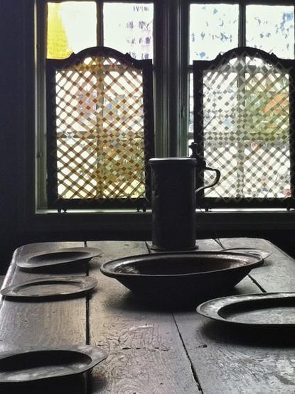 In this digital color photograph, a bright widow partly covered by latticed shades illuminates a roughly built wooden table holding pewter plates and mug, casting strong shadows.