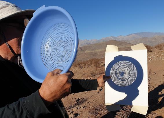 photograph of a person with their back to the sun holding a colander up to a piece of paper that is showing many partially eclipsed suns. 

This file is licensed under the Creative Commons Attribution 2.0 Generic license.

credit: 
S. Rae from Scotland, UK

source: Wikimedia Commons