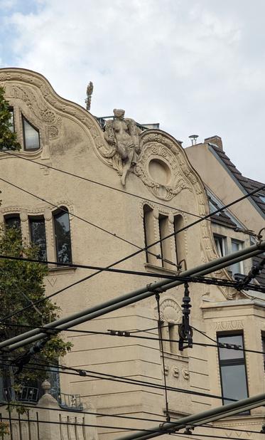 Tan colored building with a decoration of a lady near the top, probably art deco. More to the front there's a bunch of wires, some parallel, some crossing, which provide energy for the trams.