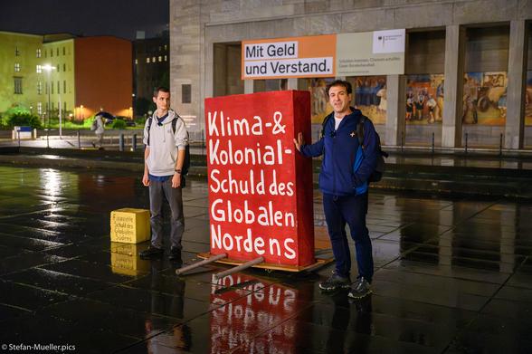 Lukas Hufert und Esteban Servat von Debt4Climate sprechen vor dem Finanzministerium zwischen zwei Betonblöcken (100kg und 1 Tonne schwer), die die Schulden des Südens und des Nordens vergleichen. Die Schulden des Nordens sind 10 Mal so groß, wie die des Südens, weshalb die Aktivist*innen einen Schuldenerlass fordern. Am Ministerium steht: „Mit Geld und Verstand. Schulden bremsen. Chancen schaffen. Unser Bundeshaushalt.“ Platz des Volksaufstands von 1953, Berlin, 12.10.2023