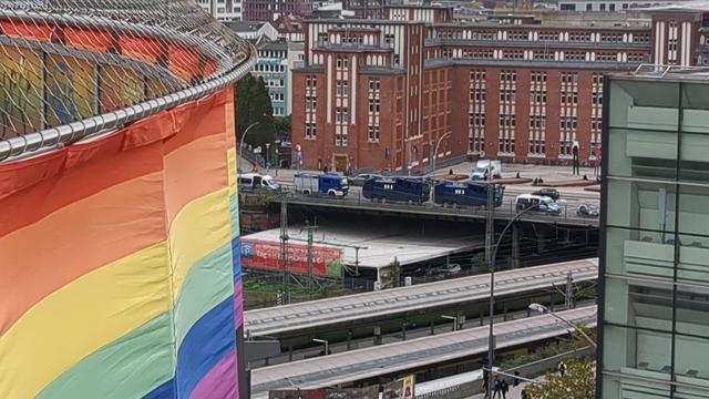 Blick vom Jupiter in Hamburg. Zwischen den Häusern auf einer Brücke über den Hauptbahnhof stehen zwei Wasserwerfer der Polizei Hamburg. Anderen Dachreling des Jupiter entlang hängt rundherum eine Regenbogenfahne.