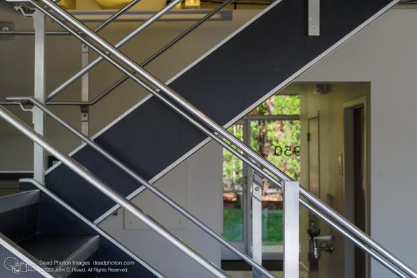 Shiny silver stairway railings criss-cross at angles in front of a black staircase. Brushed aluminum uprights support the railings. The staircase descends from the upper right and then changes direction at a landing at the lower left of the frame. A light grey wall sits behind the staircase, and at the right, a hallway leads away from the viewer past two interior doors and a drinking fountain to some glass exterior doors, through which we can see grass and trees. The number "950" appears reversed on the glass.