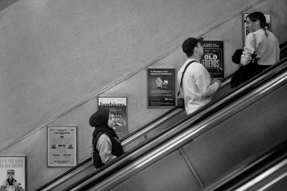 A black and white photograph of people on an escalator with a lady juxtaposed next to a poster that makes her look like the album cover from Queen’s The Miracle.