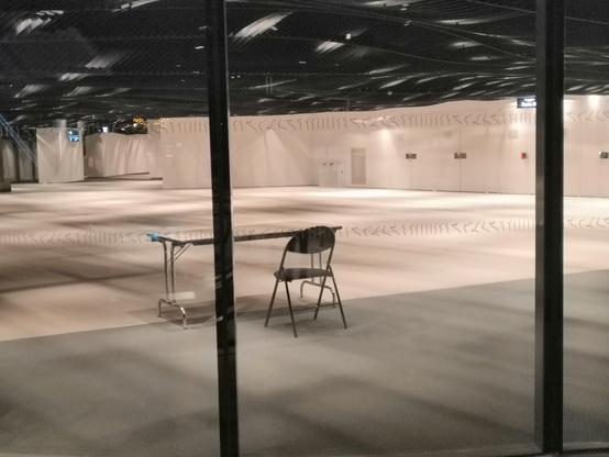 Vue d'une pièce depuis une baie vitrée. C'est un immense hall complètement vide, au bout duquel se trouvent unte table et une unique chaise.