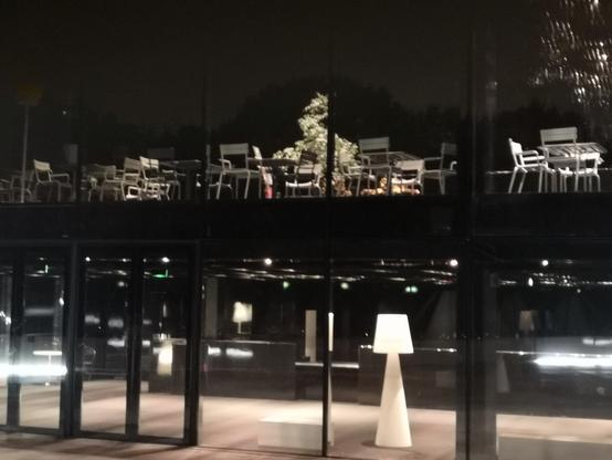 Vue de deux étages depuis fenêtres exterieurs. Au rez-de-chaussée, un hall vide dans lequel tronent des granded lampes. À l'étage, des chaises et tables de restaurant désertées mêlés à de la végétation.