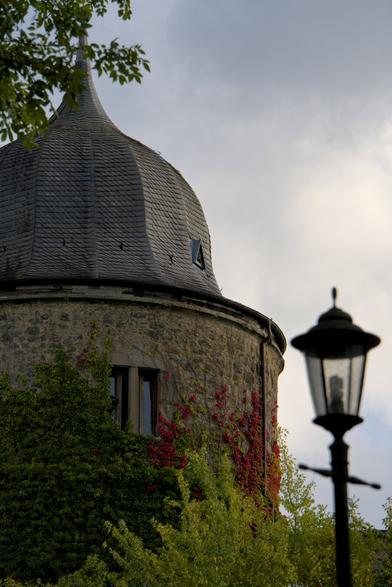 Foto de uma das torres do Castelo da Bela Adormecida em Sababurg na Alemanha. Na parte inferior há trepadeiras ainda verdes no muro, porém na torre as folhas já estão avermelhadas com a chegada do outono. A torre é cilíndrica e tem telhado em forma de domo com um espinho no topo. Na parte direita da foto em um plano mais próximo porém fora de foco, há um poste de luz pequeno com estética antiga, similar a uma lamparina antiga. O céu, visível na região superior direita da foto, está nublado.