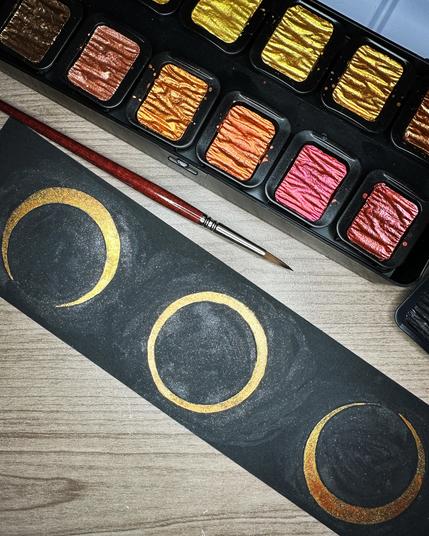 A pearlescent watercolor painting on a long black strip of paper showing three stages of today’s annular solar eclipse: two thin crescents on either side of the full “ring of fire” painted in pearlescent warm gold and rose gold tones. Around each eclipse stage is painted in glittery black.