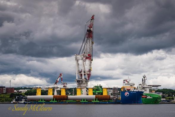 Two vessels, one with a large crane and the other with yellow windmill components , rafted up alongside each other at a wharf with an ominous sky above.
