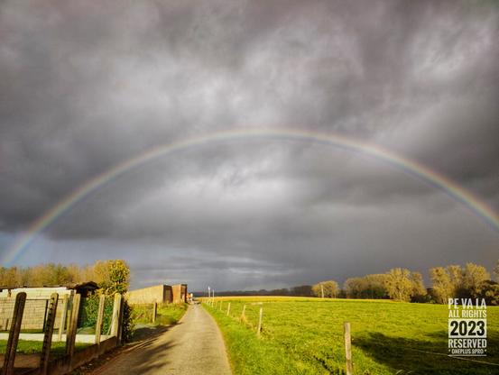 Een betonnen wegje met links tuinen, rechts weilanden. De wolken zijn donker en er is een regenboog zichtbaar.