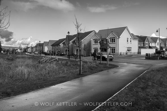 This black and white photograph shows a new residential development. There are two rows of houses at 90° angle visible in the photograph. Everything is bare and, apart from one lone person walking away from the camera in the mid distance, void of life.