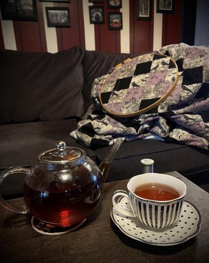 Photo for silent Sunday . Tea pot full of tea and a black and white striped teacup rest on the coffee table . A quilt in the hoop waiting to be quilted tests on the gray couch . A maroon and white striped wall of photographs behind