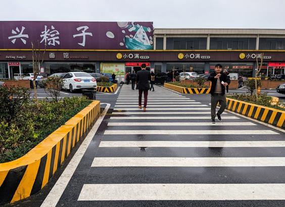 Going out from the camera is a pedestrian crossing, going through a parking lot.  On both sides of the cross-walk are knee-high barriers that are painted orange and black.  In the distance is a two-story building that stretches the width of the image.