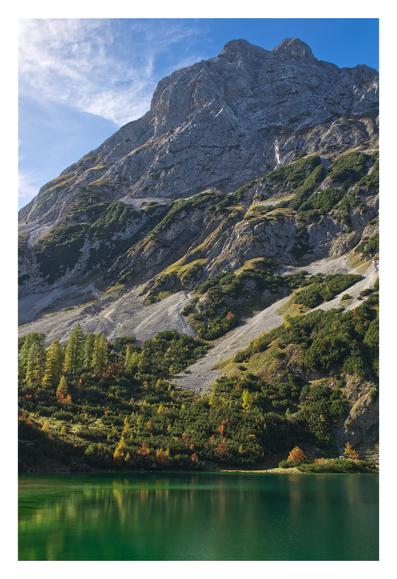 View of Sonnenspitze from its base on the other shore of Seebensee. Patches of colorful trees (larch, mountain pines) covering the lower parts of the talus fields. Lush, deep green water color (from this POV) and almost still lake surface with soft reflections. Bright blue sky with some (still faint) clouds forming.