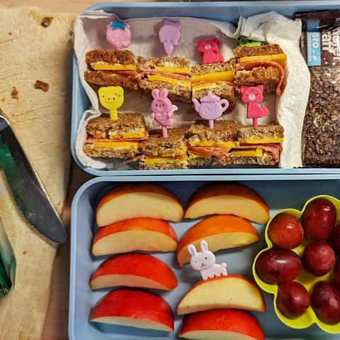 Draufblick auf die 2 Ebenen einer himmelblauen, rechteckigen Bentobox. In der oberen Schale liegen kleine Canapés aus dünnem Vollkornbrot mit Butter-, Salami- & Cheddar-Belag. In ihnen stecken kleine Plastikspieße mit niedlichen Tierchen und Gegenständen (Häschen, Eistüte, Erdbeere usw.) in vorrangig Pinktönen. Rechts daneben liegt ein Schoko-Müsliriegel ohne Zucker. In der unteren Dose liegen links Apfelspalten, in einer Spalte steckt ein dreizackiger, weißer, kleiner Plastikspieß mit Hasenmotiv als Griff. Rechts davon ist eine Silikonmuffinform in Form einer Blüte. In jedem Blütensegment steckt eine rote Weintraube.
Links ist ein bekrümeltes Holzbrettchen und ein benutztes Messer neben den Boxen.