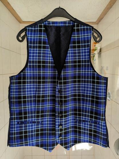 eine blau karierte Weste mit dünnen weißen Streifen und schwarzen Flächen dazwischen hängt auf einem Kleiderbügel