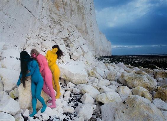 painted photo: a blue, a pink, and a yellow woman shimmy close about some white rocks