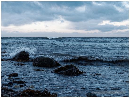 Das Foto zeigt die Ostsee zur Blauen Stunde in der Abenddämmerung unter einem locker bewölkten, leicht rosa angehauchten Himmel, im Vordergrund liegen eiszeitliche Findlinge in der Brandung.