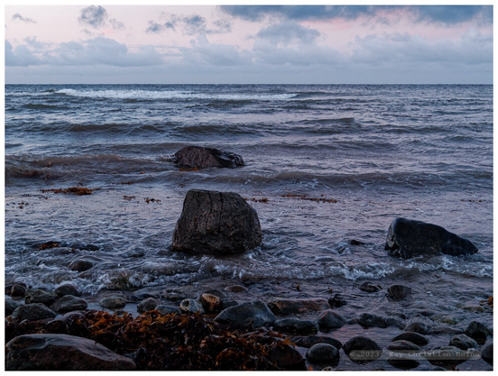 Das Foto zeigt die Ostsee zur Blauen Stunde in der Abenddämmerung unter einem locker bewölkten, leicht rosa angehauchten Himmel, im Vordergrund liegen eiszeitliche Findlinge in der Brandung, ganz vorn ist der von grobem Schotter bedeckte Spülsaum des Strandes zu sehen.
