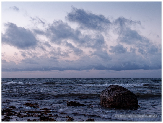 Das Foto zeigt die Ostsee zur Blauen Stunde in der Abenddämmerung unter einem locker bewölkten, leicht rosa angehauchten Himmel, im Vordergrund liegen eiszeitliche Findlinge in der Brandung.