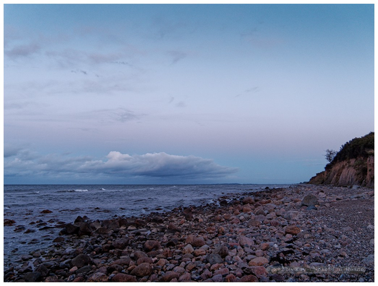 Das Foto zeigt eine Weitwinkelaufnahme des von grobem Schotter bedeckten Ostseestrandes zur Blauen Stunde in der Abenddämmerung unter einem locker bewölkten, leicht rosa angehauchten Himmel. Die Linien des Horizonts und Spülsaums sowie des Steilufers am linken Bildrand führen auf den Fluchtpunkt im unteren rechten Drittel des Fotos hin.