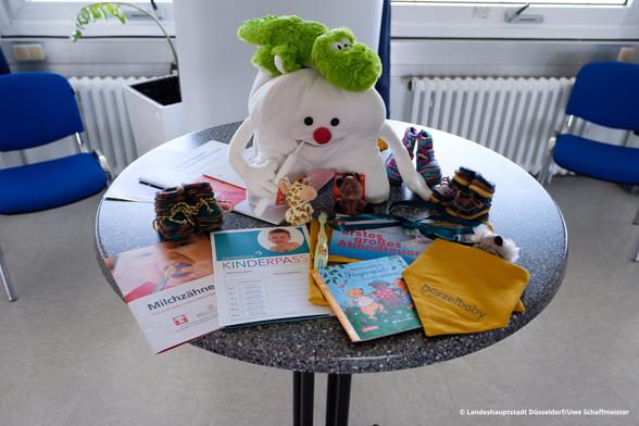 Ein Tisch mit dem Inhalt des Willkommenspakets für Eltern von Neugeborenen, welches der Elternbesuchsdienst ausgibt. Viele Broschühren, zwei Kuscheltiere (ein Zahn, ein Dino), Socken, Schühchen, ein "düsselbaby" Lätzchen und einiges anderes mehr.