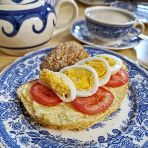 Esstisch aus hellem Holz. Im Fokus ein weißer Teller mit blauem, floralem Motiv (Serie "Burgenland" von Villeroy & Boch). Auf dem Teller eine Schreibe Weißbrot, bestrichen mit Butter, darauf eine Lage dünne Scheiben Tomate, dann fächerartig ausgebreitet Scheiben von hartgekochtem Ei, darauf ein paar Salzkristalle. Daneben liegt der Brotkanten auf dem Teller. Im Hintergrund rechts eine Teetasse der gleichen Serie gefüllt mit Milchkaffee. Links eine kleine Teekanne in weiß mit großem, blauem Wellenmuster.